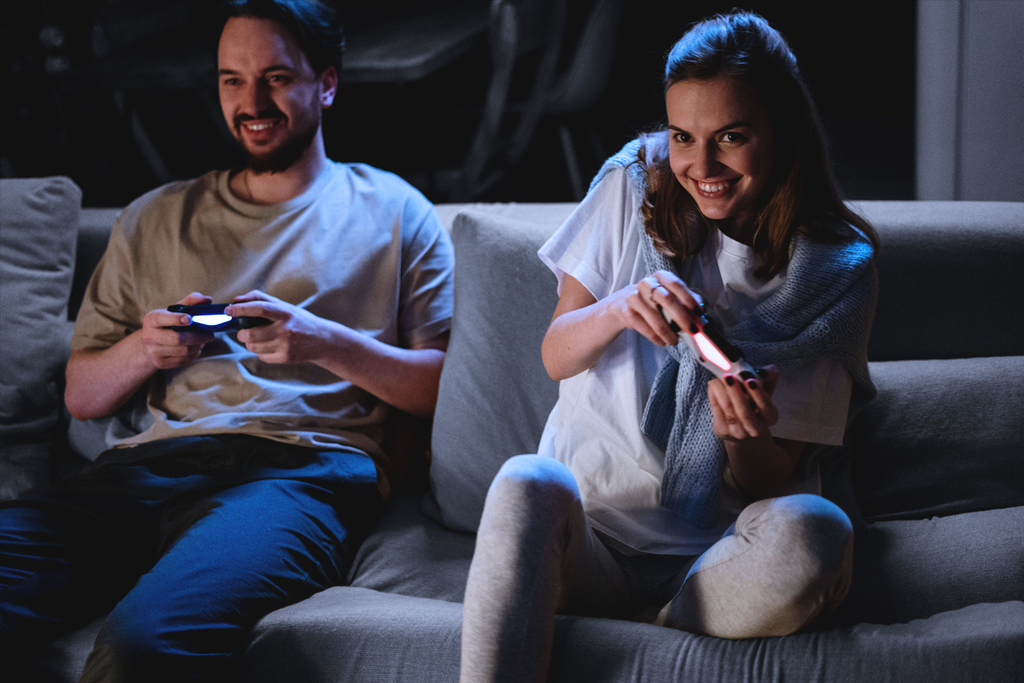 Zu sehen sind zwei Personen auf einem Sofa, die Videospielcontroller halten und scheinbar ein Spiel spielen
